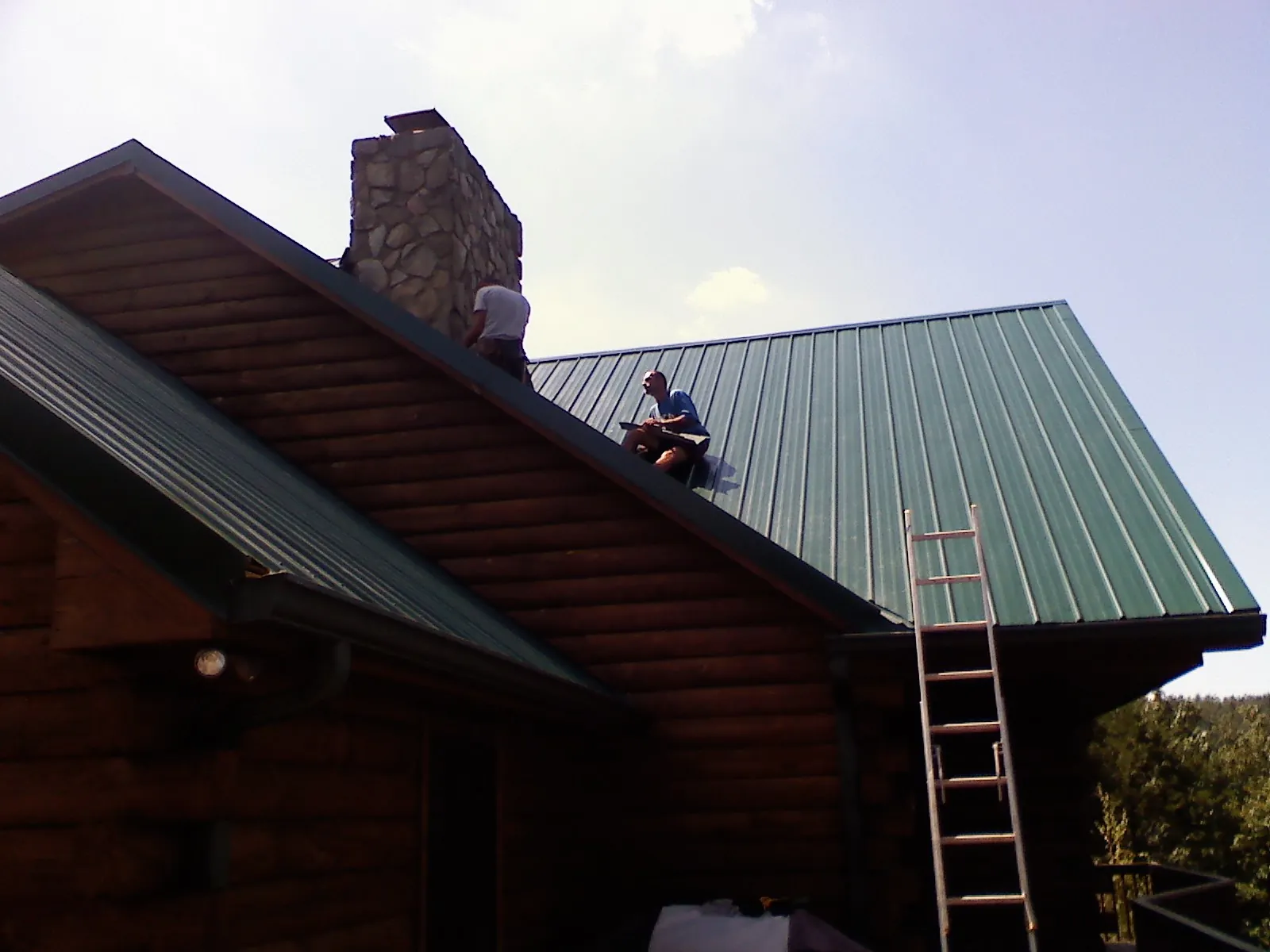 Roofers installing green metal panels beside a stone chimney on a log home in South Bend